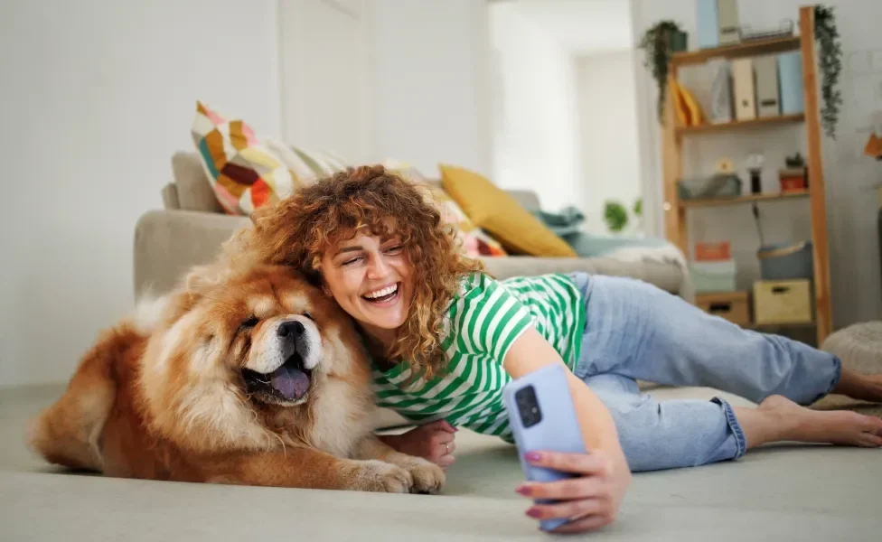 Tutora tirando foto em celular ao lado de cachorro Chow Chow 